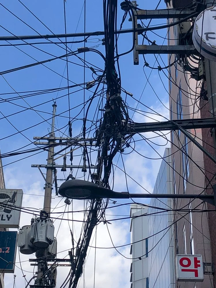 A tangled web of electrical wires and cables hanging between buildings, with a streetlamp and utility poles visible against a partly cloudy sky.