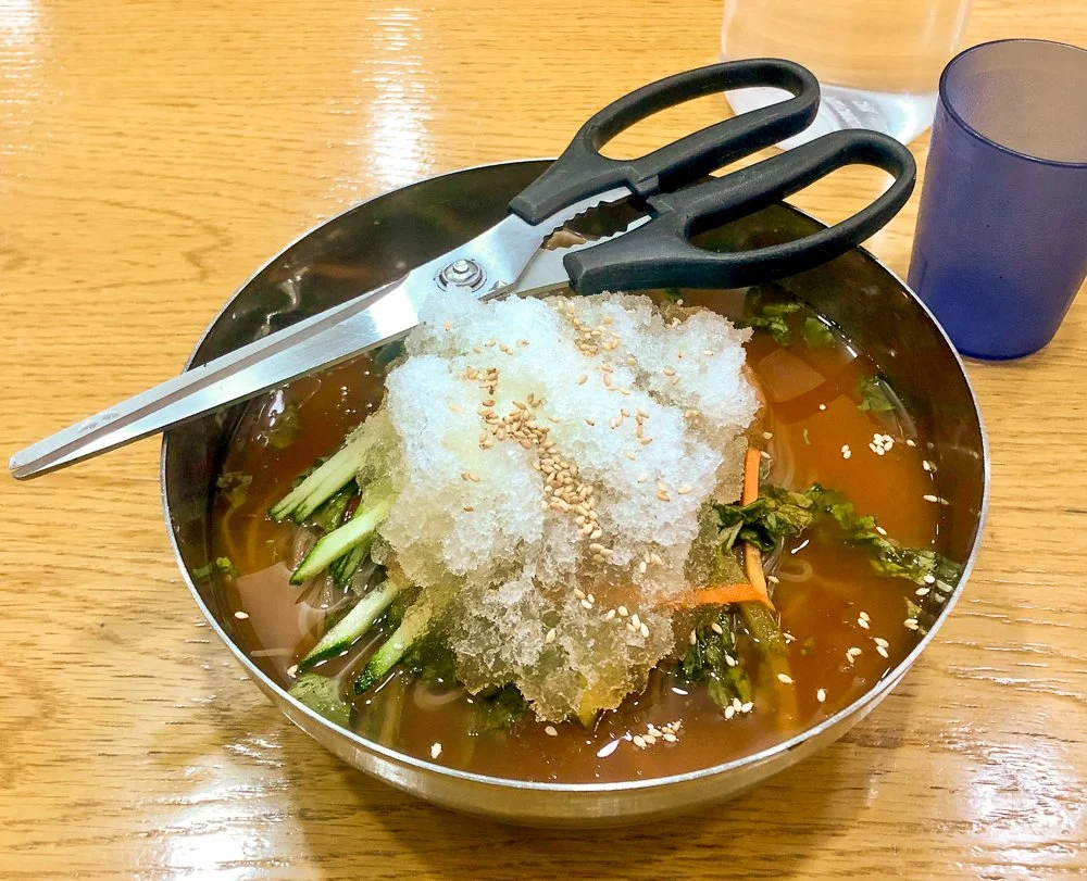 Bowl of Asian cold noodle soup with shredded vegetables and crushed ice, next to scissors and a glass of drink.
