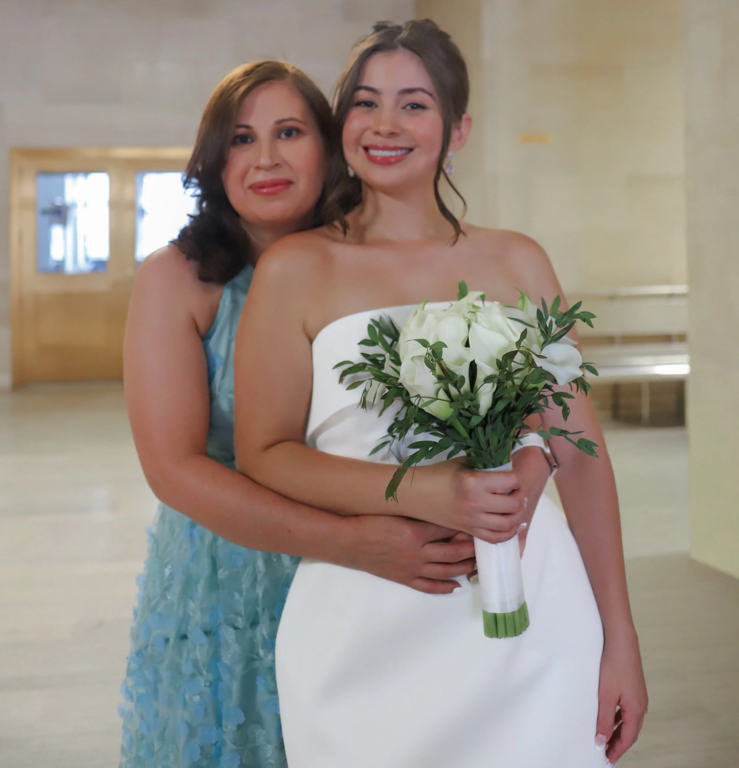 A bride holding a bouquet of white flowers, with another woman hugging her from behind, in a bright indoor setting.