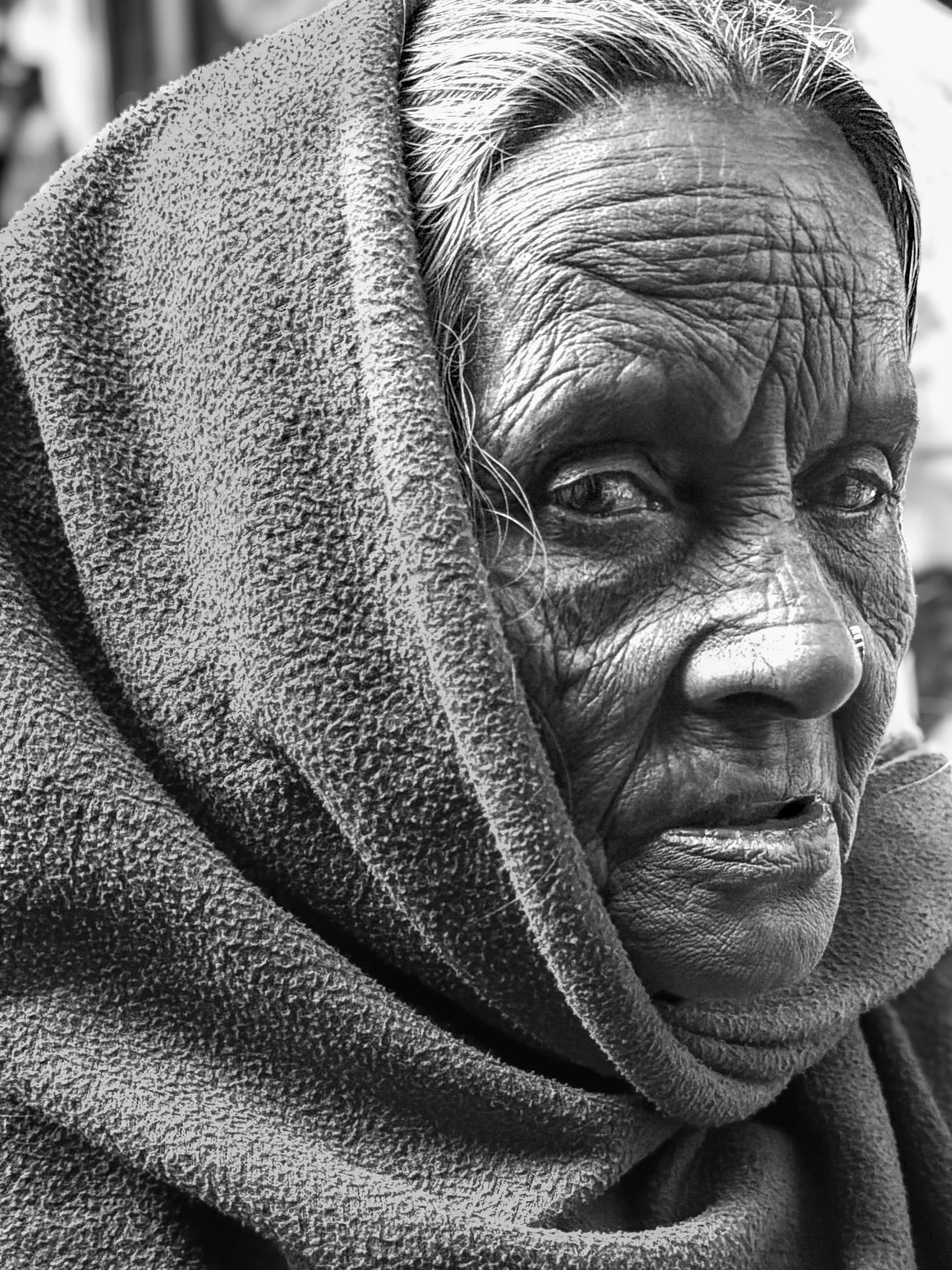 Close-up black and white photograph of an elderly woman with a textured shawl over her head, showing detailed facial features and deep wrinkles.