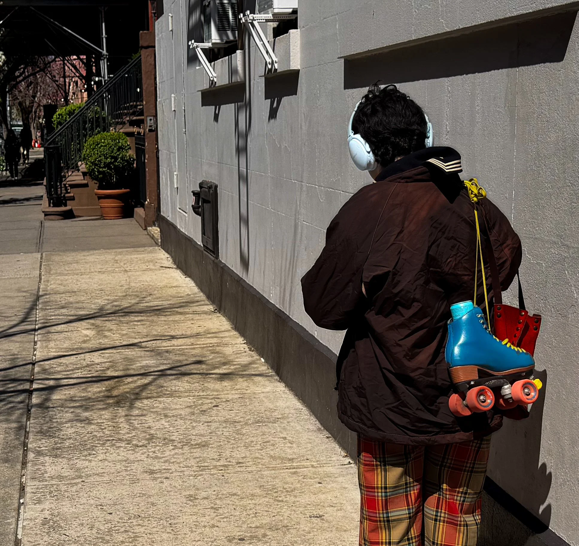 Person wearing a dark jacket, plaid pants, and white headphones standing on a sidewalk with a gray wall in the background, carrying blue roller skates and a red shoe.
