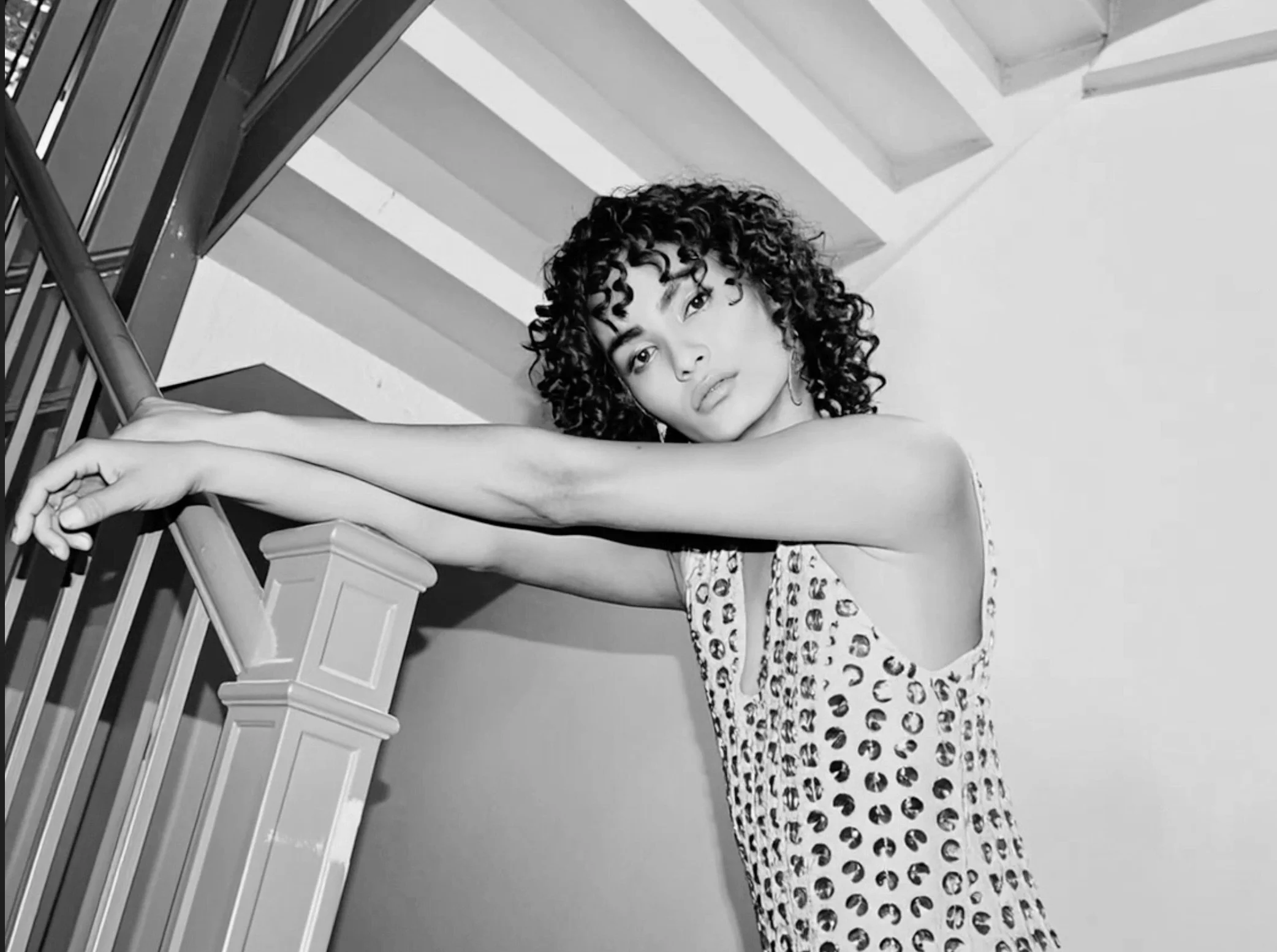Black and white photo of a woman with curly hair leaning on a staircase railing, looking at the camera with a serious expression, wearing a sleeveless patterned top.