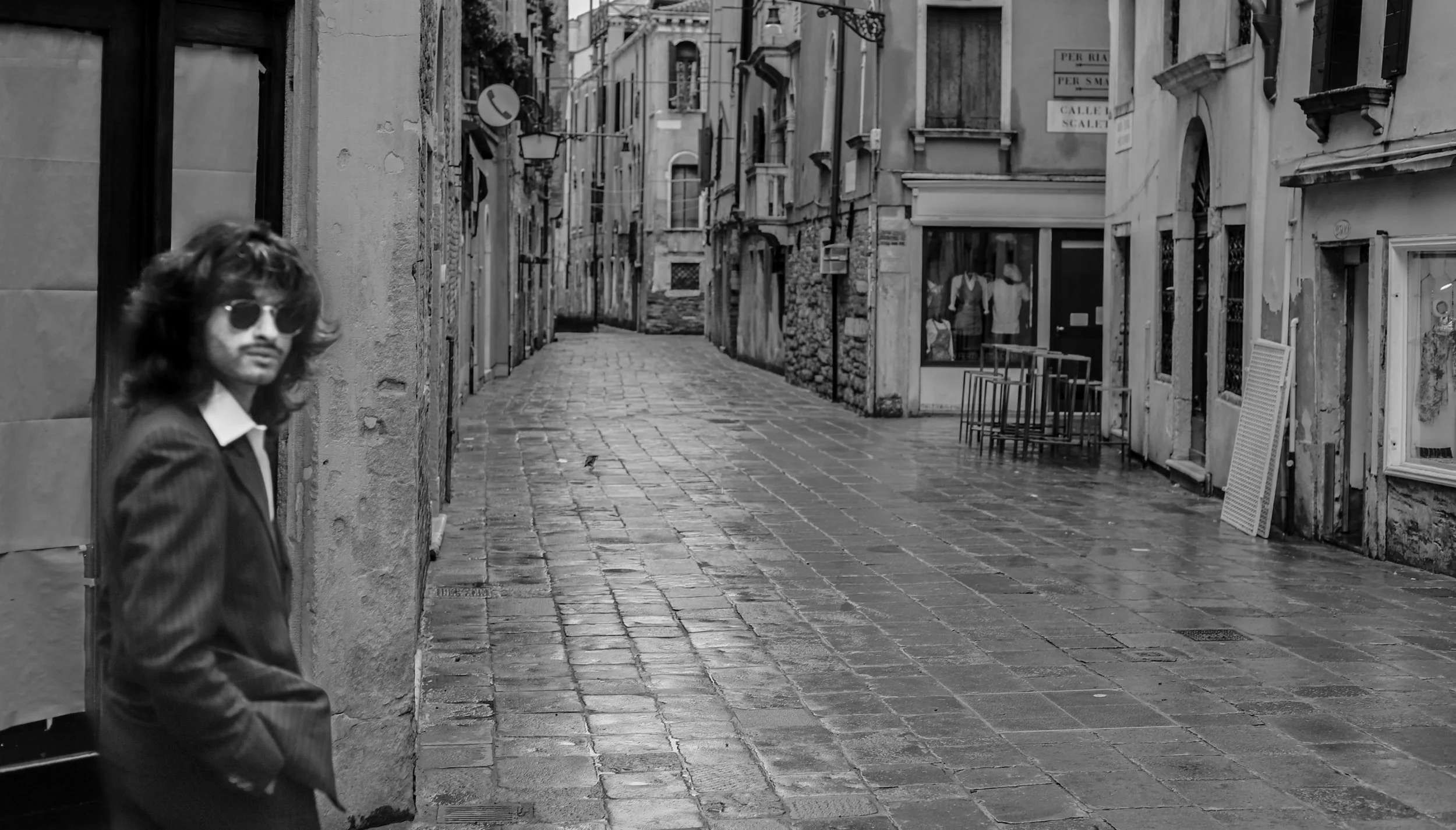 A man with long hair, wearing sunglasses and a suit, leaning against a wall on a wet, cobblestone street in a European city.
