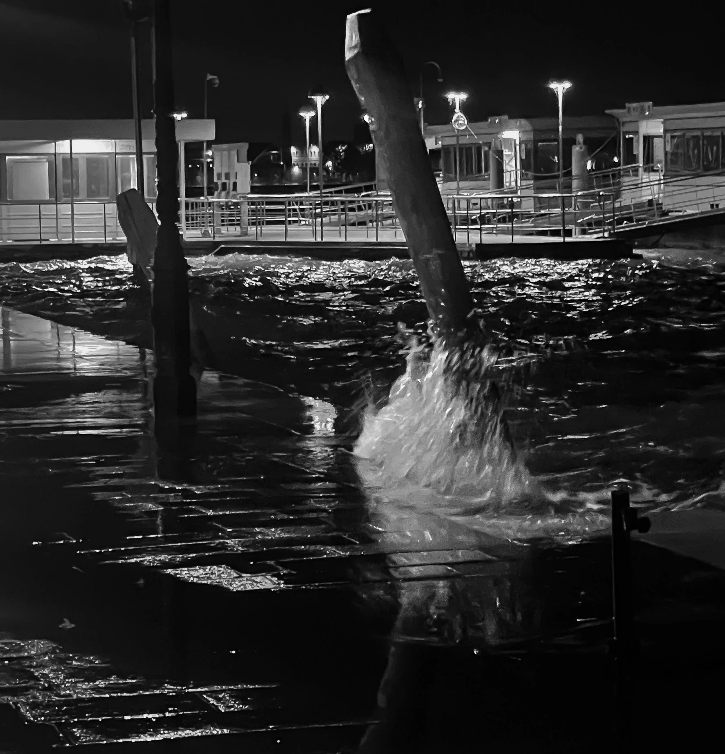 A whale's tail splashing as it dives into the water at night near a dock with boats and lights.