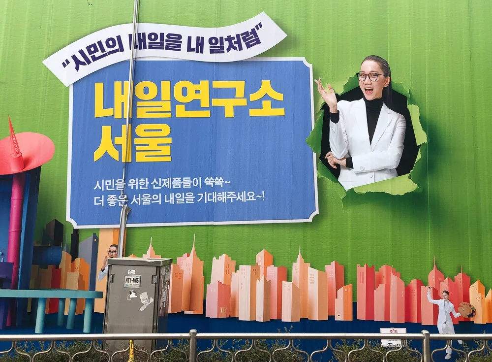 Large green billboard with South Korean text, a woman in a white coat emerging from a torn paper hole, a cityscape illustration, and small cutouts of people in lab coats.