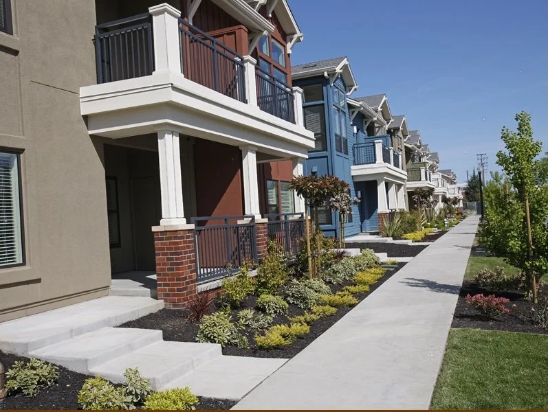 A row of new multi-story houses with small front yards and a sidewalk in front, under a clear blue sky.