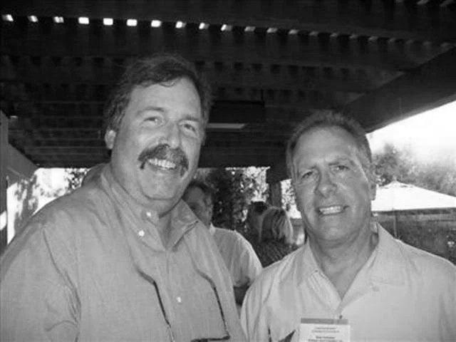 Two men smiling and posing for a photo outdoors under a wooden roof, with other people in the background.