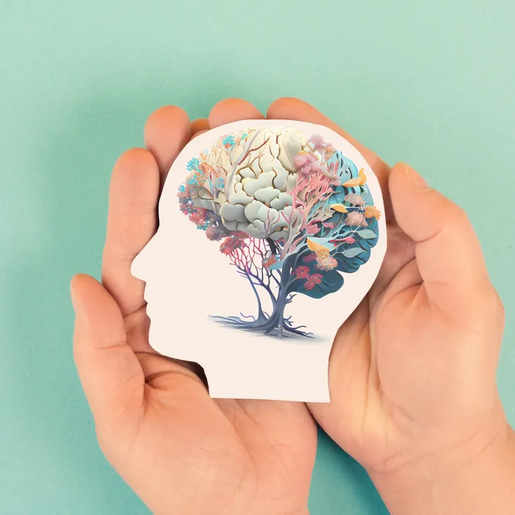 A hand holding a paper cutout of a human head silhouette with a tree and colorful abstract elements inside, symbolizing the mind or thoughts.