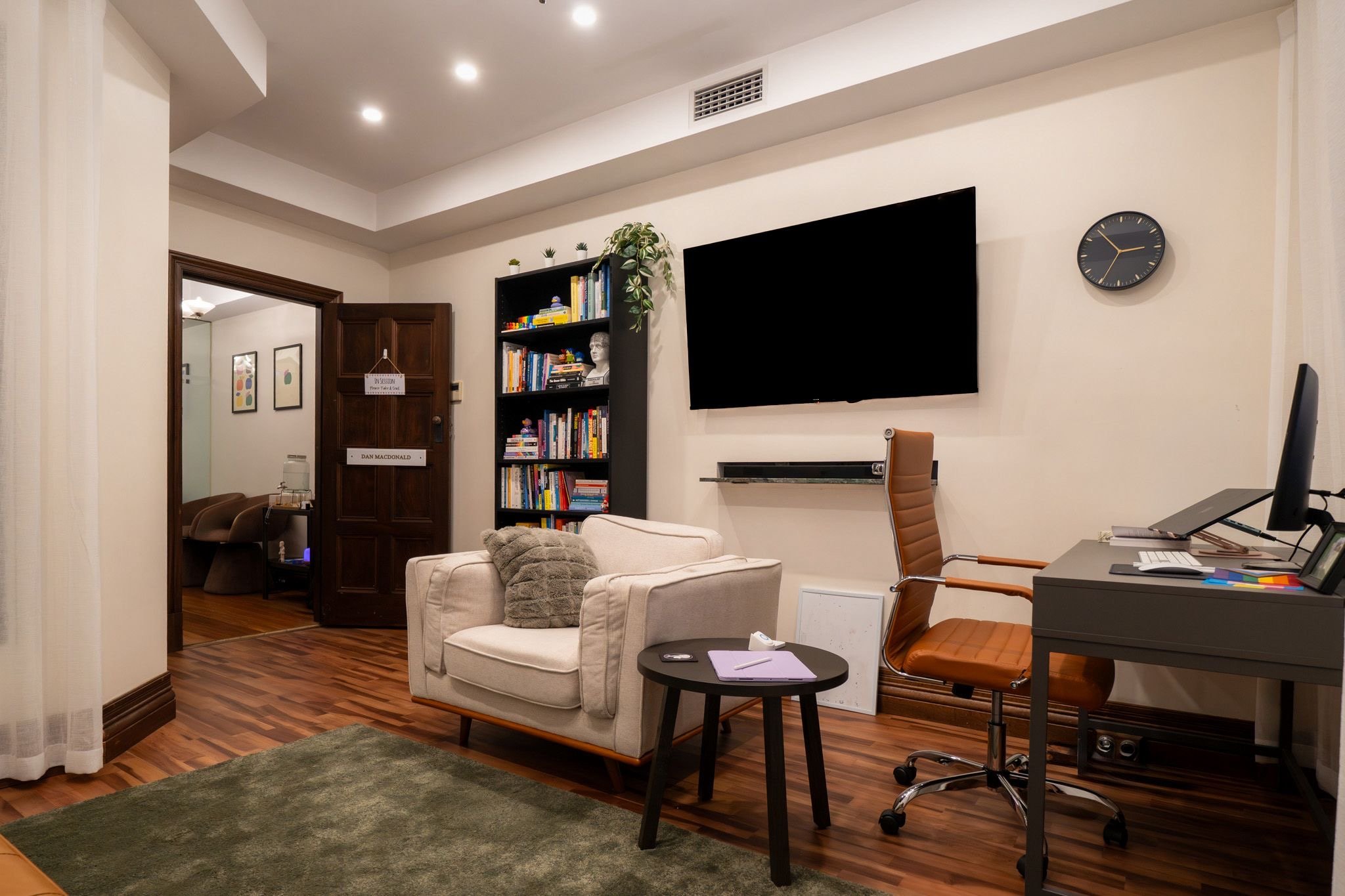 Living room with a beige armchair, a black bookshelf filled with books, a large wall-mounted TV, a brown office chair, a small black side table with papers, and a black desk with a computer, lamp, and files. Wooden flooring, a green rug, and a wall clock are also visible.