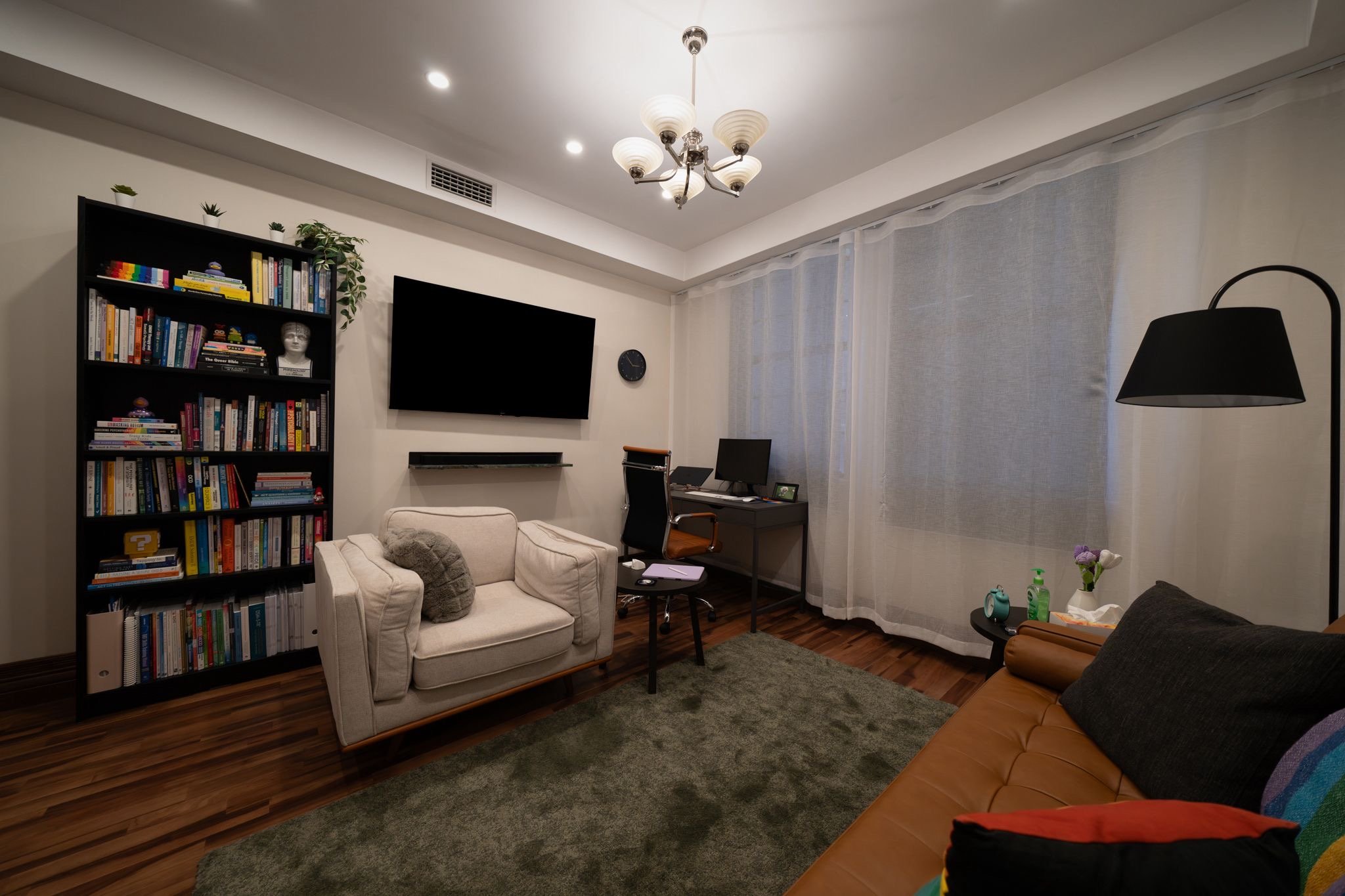 A living room with white walls, hardwood floors, a flat-screen TV mounted on the wall, a beige armchair with a pillow, a black bookshelf filled with books and decorative items, a small black side table, a brown leather sofa with pillows, a floor lamp, a window with sheer curtains, and various small decorative items on furniture.
