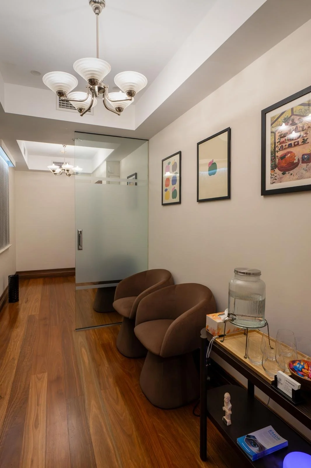 Interior of a modern room with two brown chairs, a water dispenser on a side table, framed artwork on the wall, and wooden flooring.