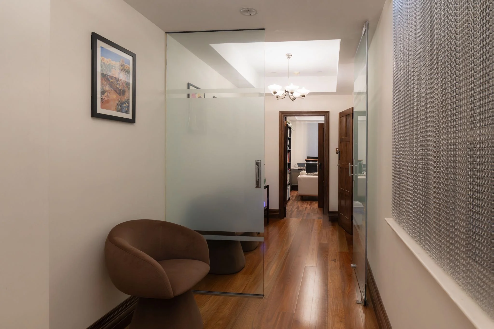 A hallway with a wood floor, white walls, and a leather chair on the left. A frosted glass door is on the left, and a wooden door on the right. A painting hangs on the wall to the left, and a chandelier hangs from the ceiling in the background. The hallway leads to a room with a white sofa and bookshelf.