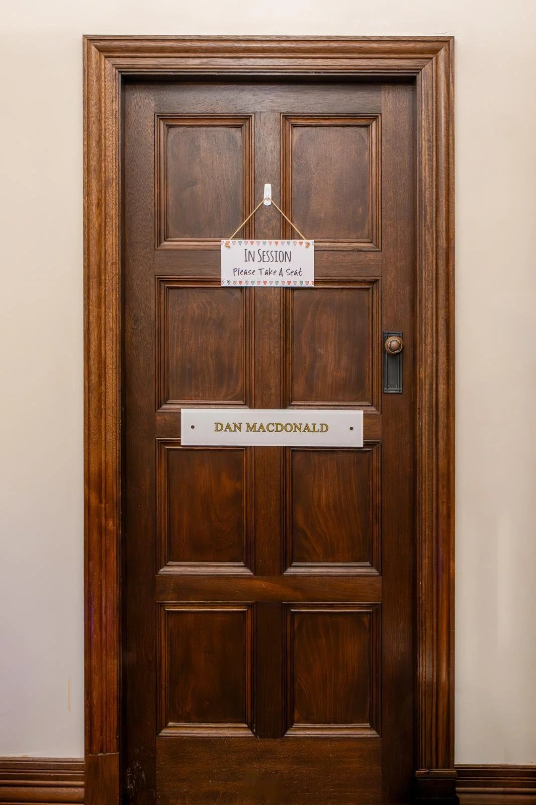 A wooden door with a sign that reads 'In Session Please Take a Seat' hanging from a hook, and a nameplate that says 'Dan Macdonald'.