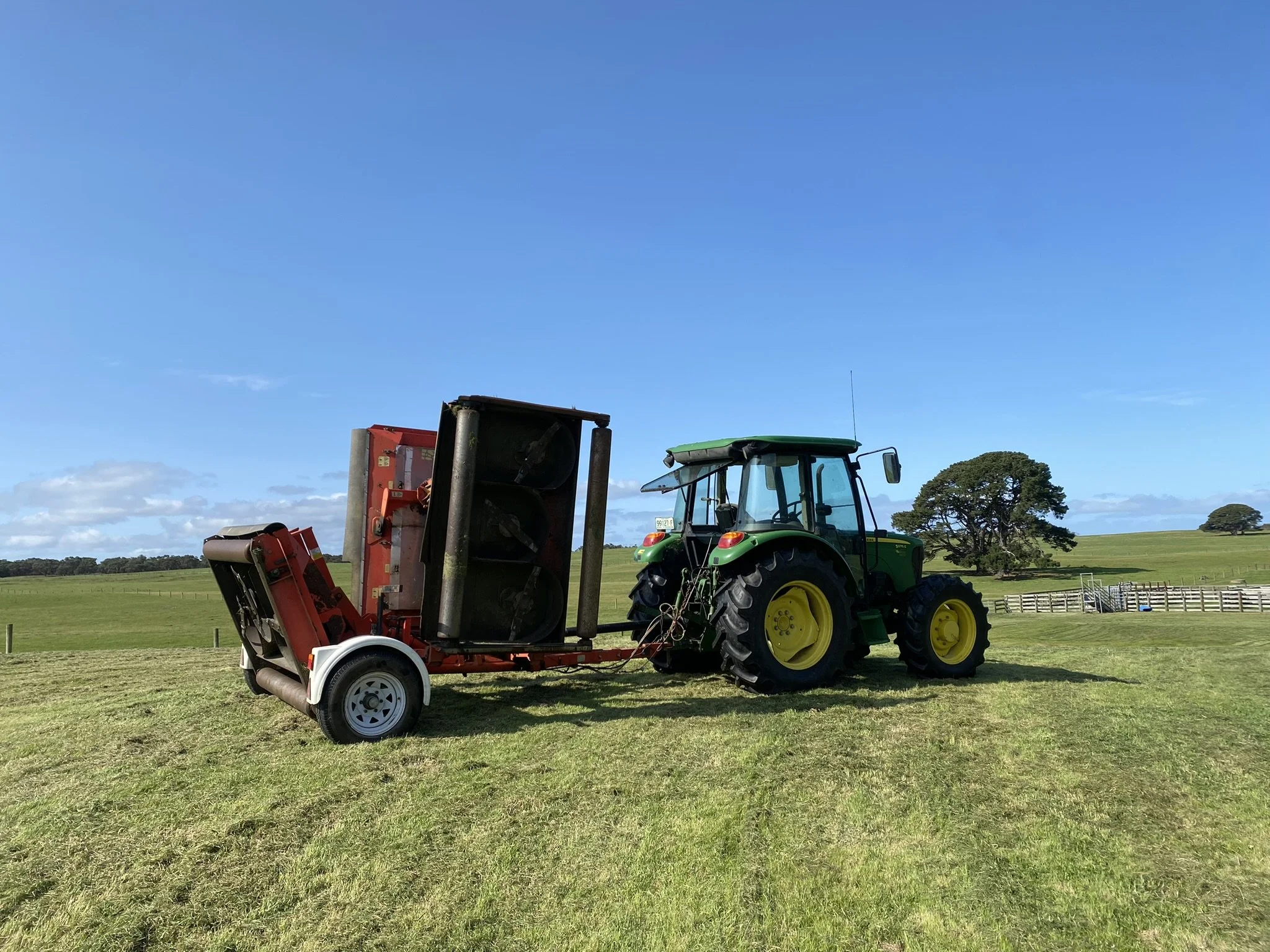 Green tractor attached to a red lawn mower on a vast grassy field under a blue sky with scattered clouds, trees in the background.