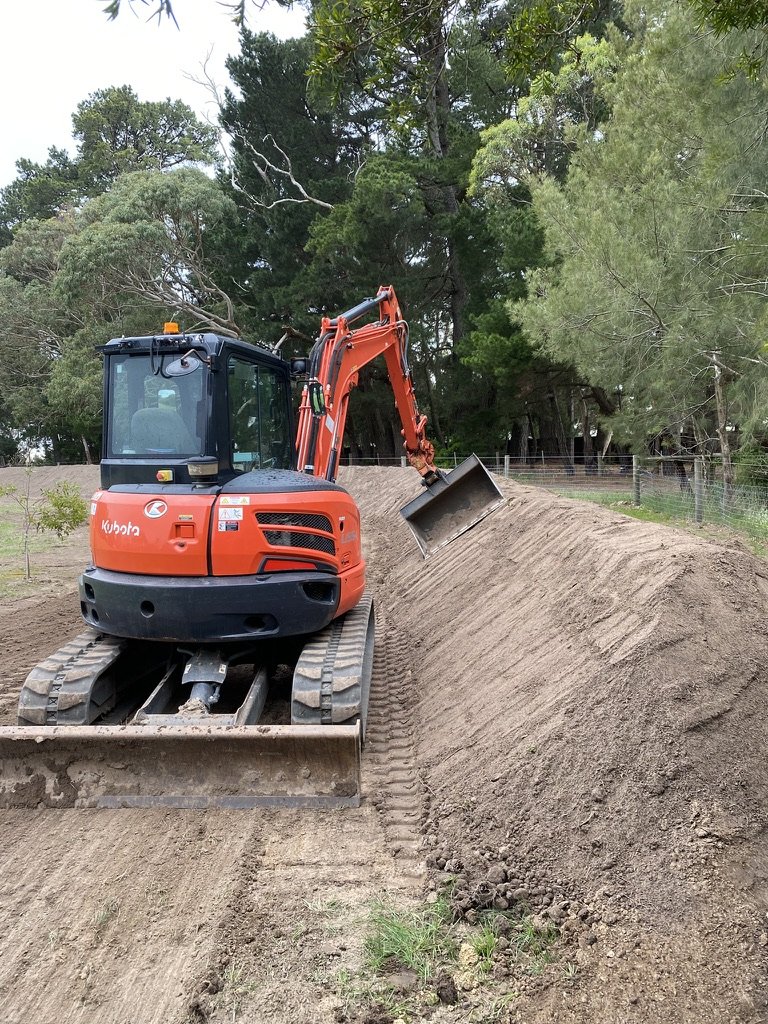 A small orange excavator moving dirt on a sloped construction site, surrounded by trees.