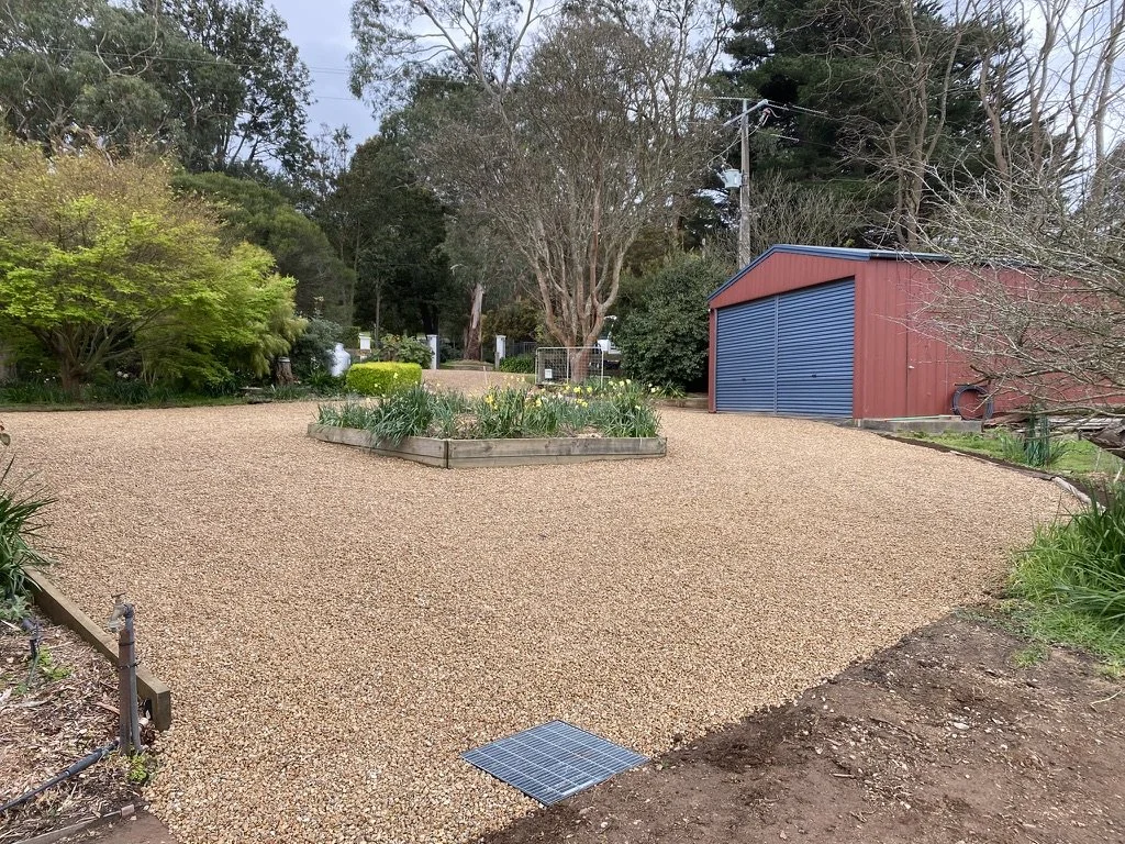 A landscaped yard with a gravel driveway, a flower bed in the center, green bushes, trees, and a red and blue shed.