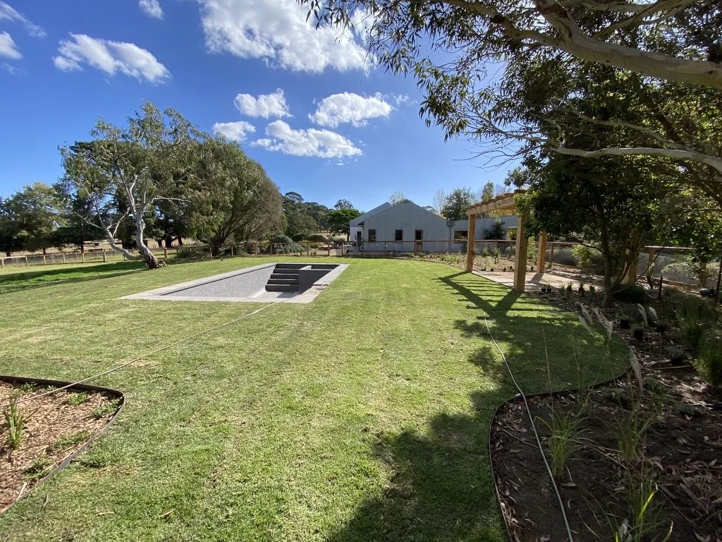 A spacious backyard with a grassy lawn, trees, a small concrete pool with stairs, a garden bed on the right, and a barn-like building in the background, under a partly cloudy sky.