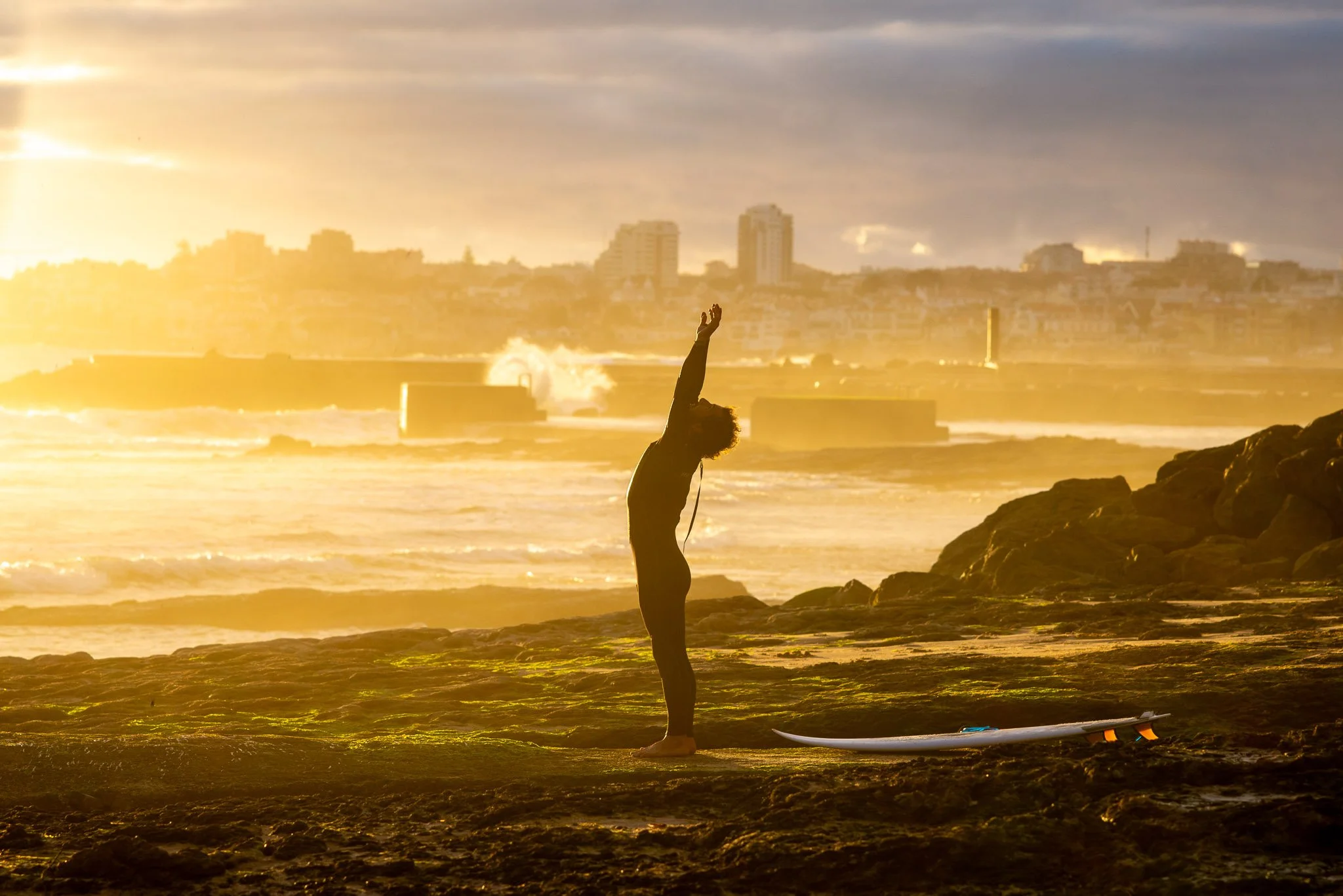 Person doing yoga on rocks at the beach during sunset, with a surfboard lying nearby and city buildings in the background.
