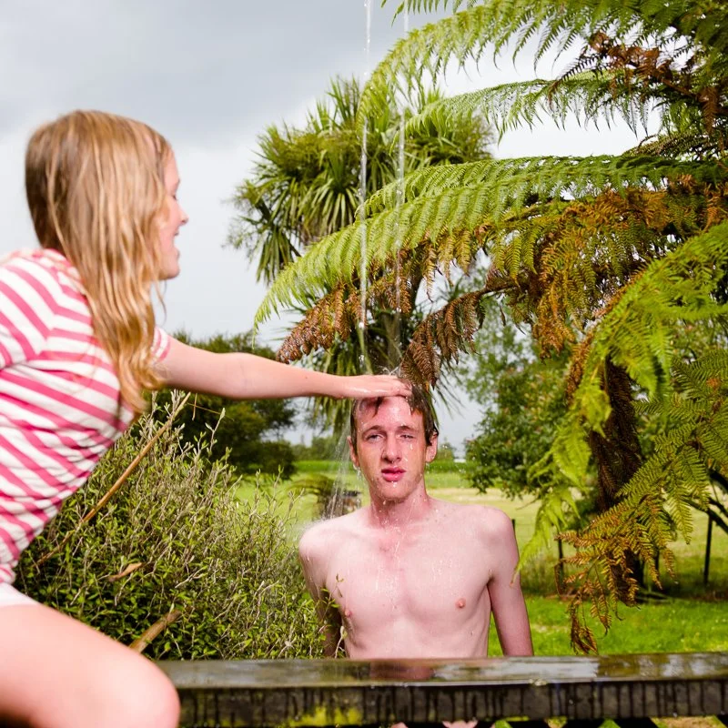 A woman in a pink and white striped shirt offers water to a shirtless man under a fern with water pouring over his head outdoors in a lush green garden.