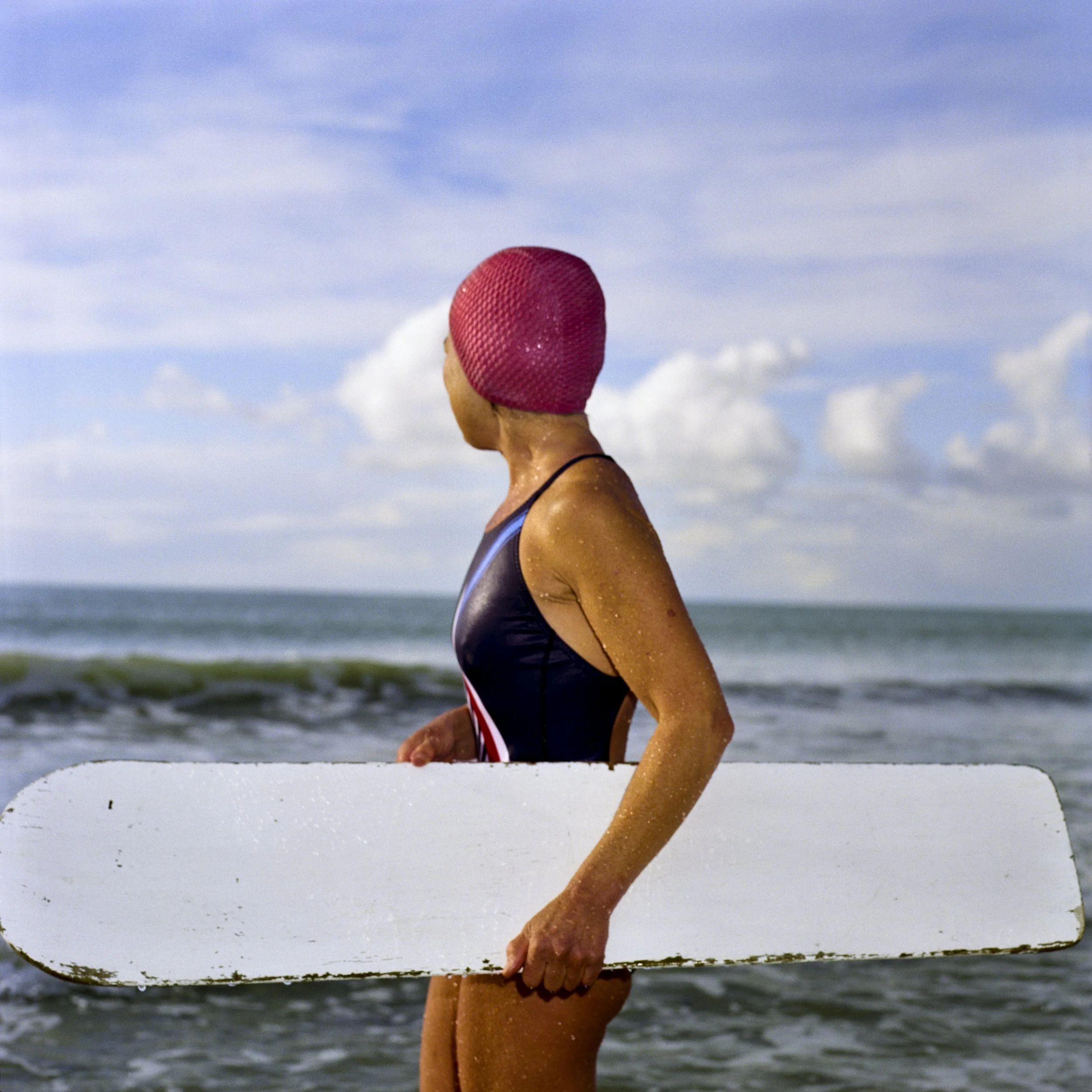 Woman in swimsuit holding a white paddle at the beach, wearing a pink swim cap, with ocean and cloudy sky in the background.