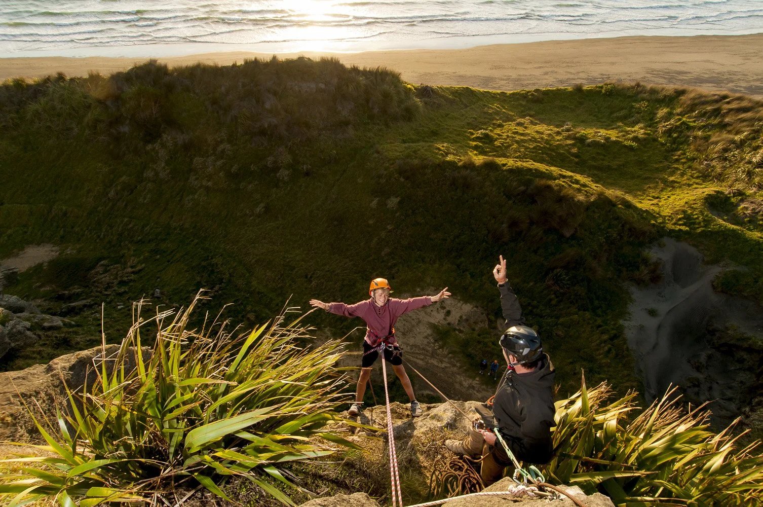 Two people with helmets and climbing gear on a rocky cliff, with one person reaching out and smiling, and the other raising a hand in a gesture, overlooking a green valley and a beach in the background at sunset.