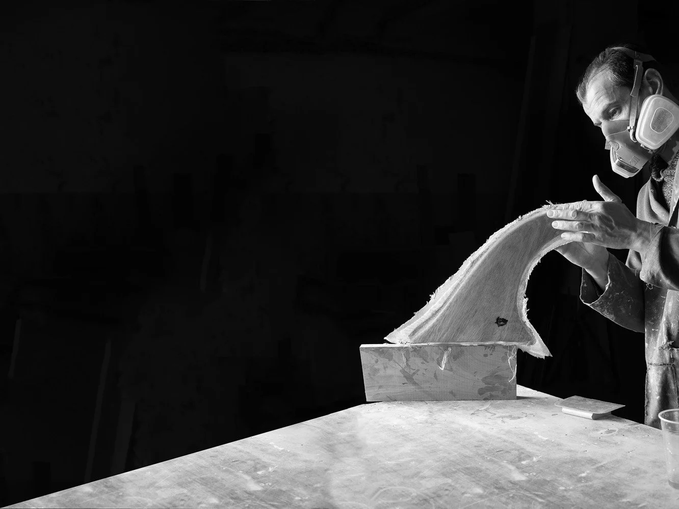 A man wearing a mask and protective goggles shaping a piece of wood, possibly woodworking or sculpting, in a workshop.