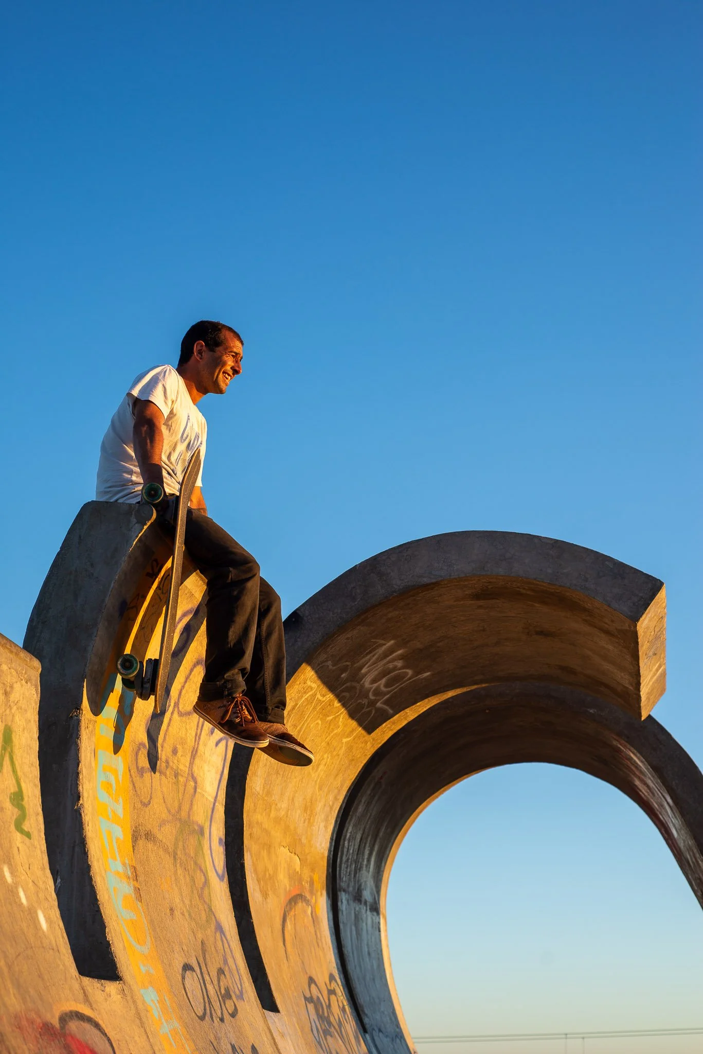 A man sitting on the edge of a skate park bowl, holding a skateboard, during sunset with a clear blue sky in the background.
