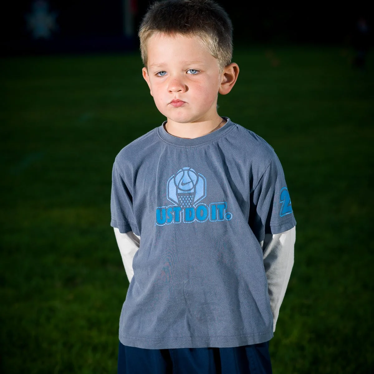 Young boy wearing a gray basketball jersey with the phrase "JUST DO IT" and a basketball graphic on the front, standing outdoors on a grassy field at night.