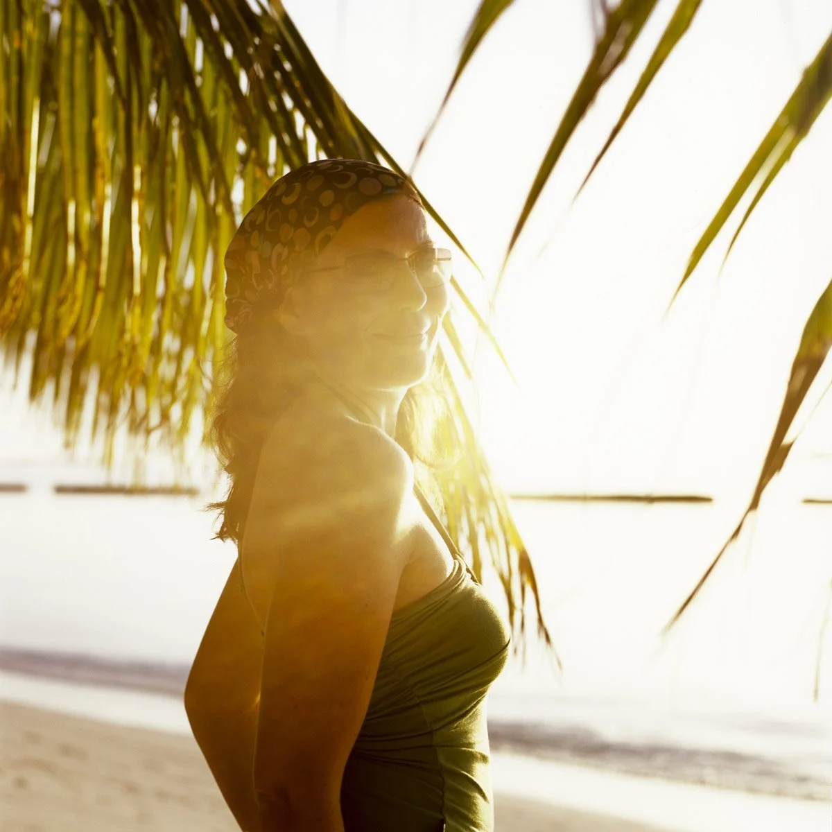A woman standing under palm trees on a beach during sunset, smiling, wearing glasses, and a headscarf.