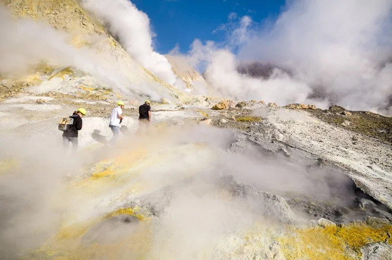 Three people exploring a volcano site with steam rising from the ground, surrounded by rocky terrain and bright blue sky.