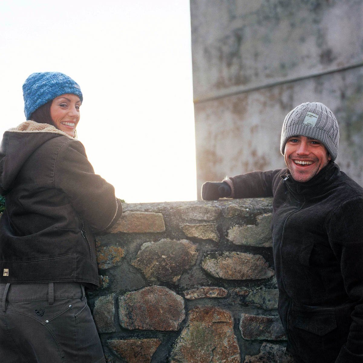 A woman and a man wearing winter clothing and knit hats, smiling while leaning on a stone wall outdoors.