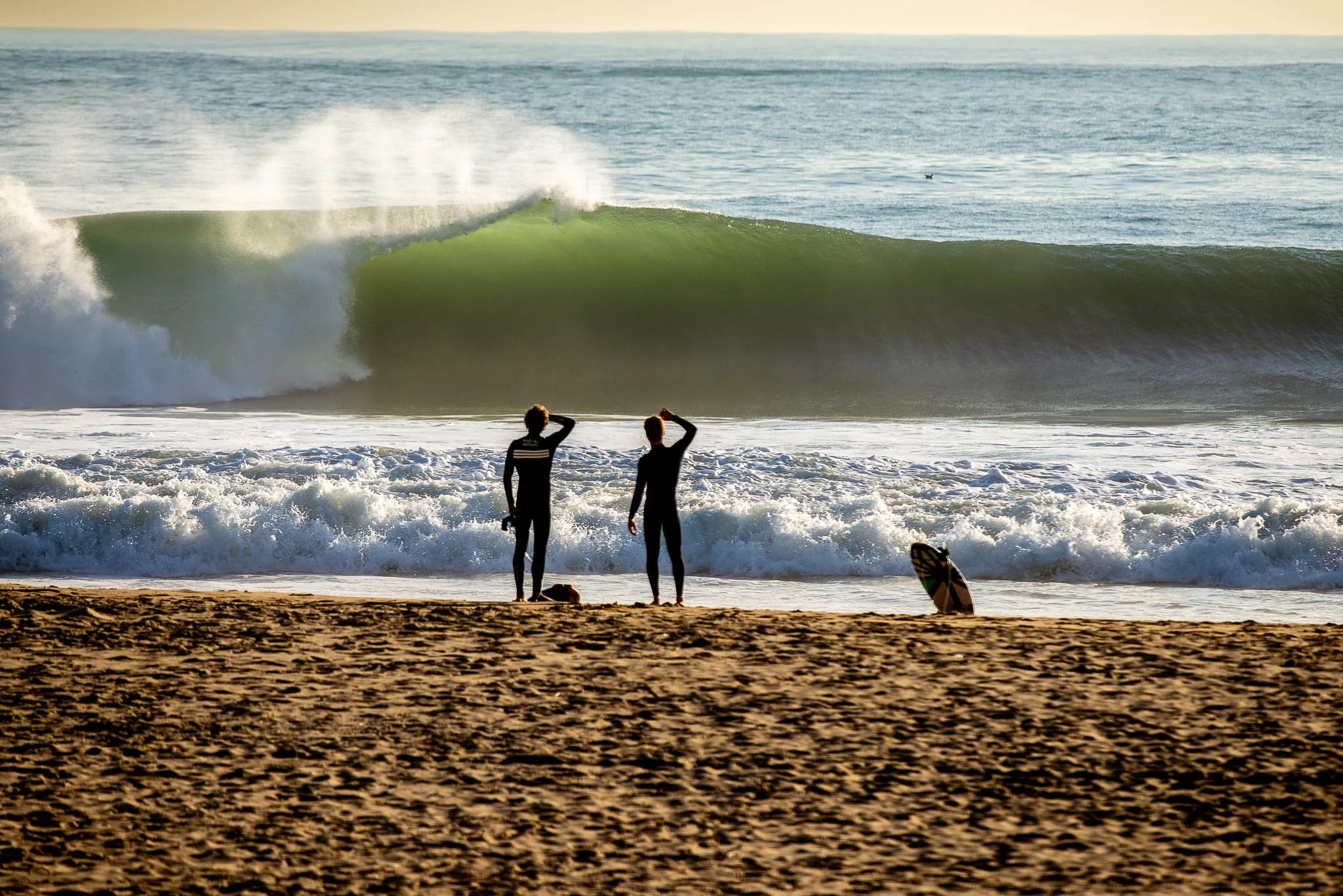 Two surfers in wetsuits standing on the sandy beach, looking out at the large ocean waves during sunset.