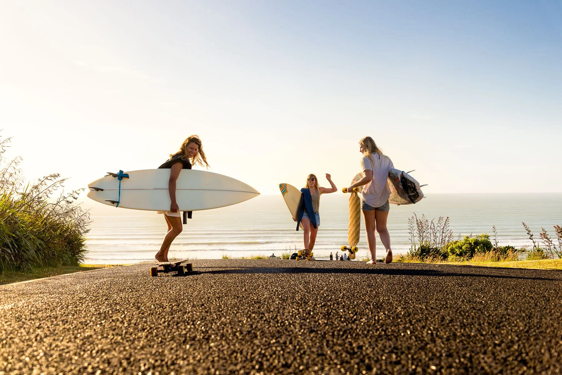 Three young women with surfboards and skateboards near the beach during sunset.