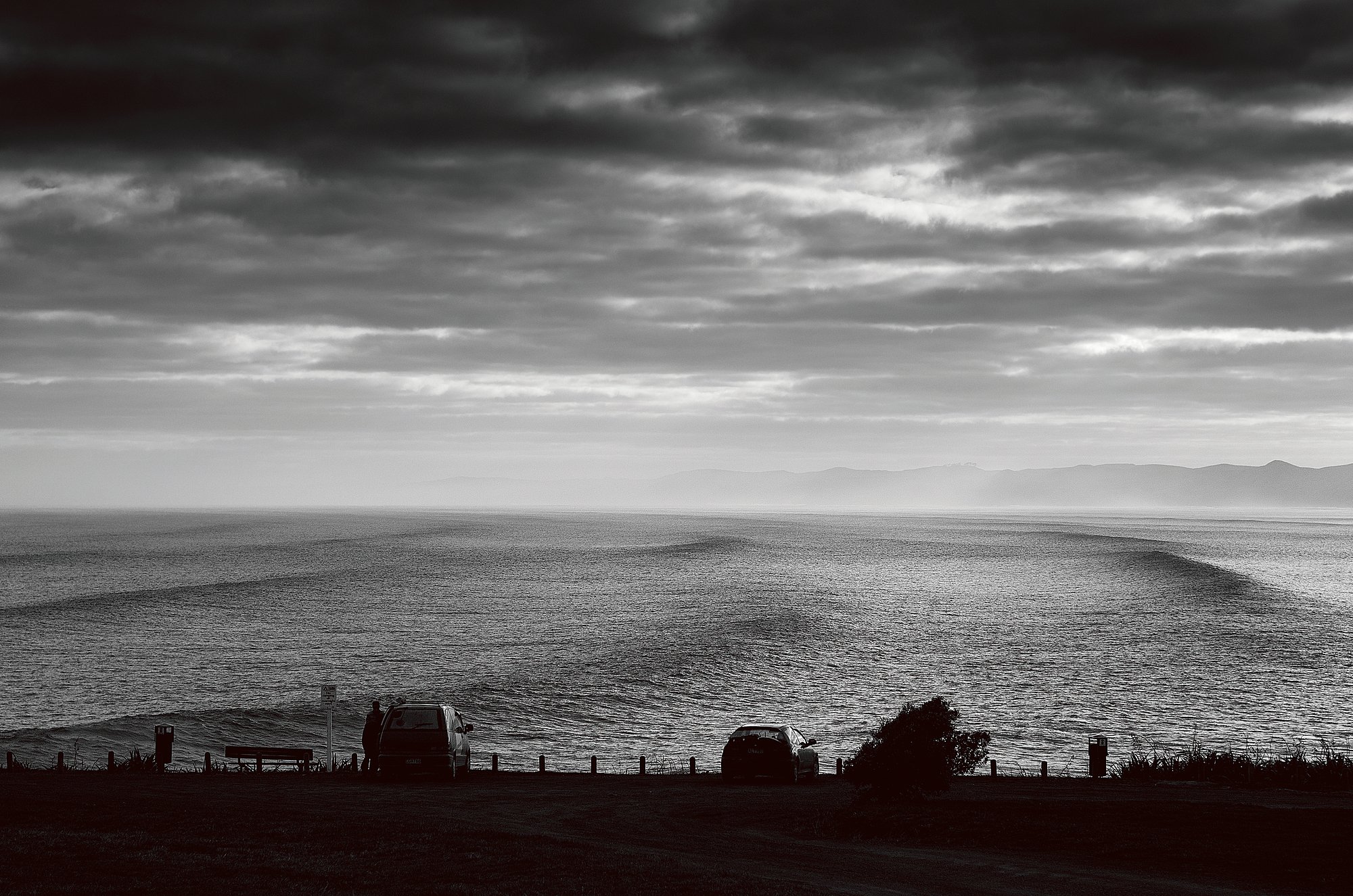 A study of the morning arrival at Manu Bay. In the high-contrast light of the Tasman Sea, spectators and surfers gather at the lookout to survey the lines. It captures the distinct split-second of the Raglan ritual: the quiet observation from the car