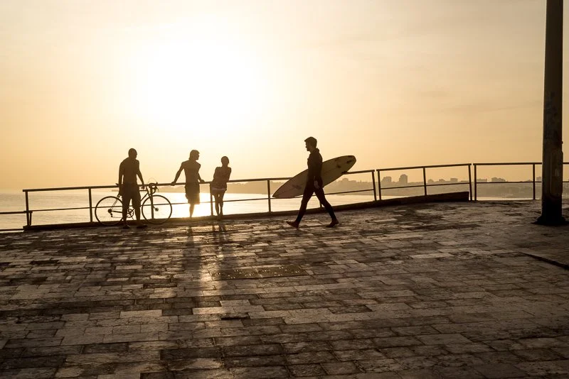 Silhouetted people at the beach during sunset, with one person carrying a surfboard and another with a bicycle.