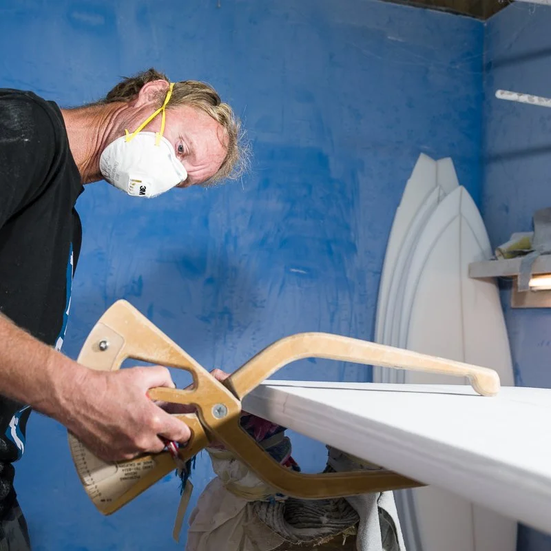 A man wearing a white face mask with yellow straps, working with a saw on a surfboard in a workshop with blue walls and surfboard fins in the background.