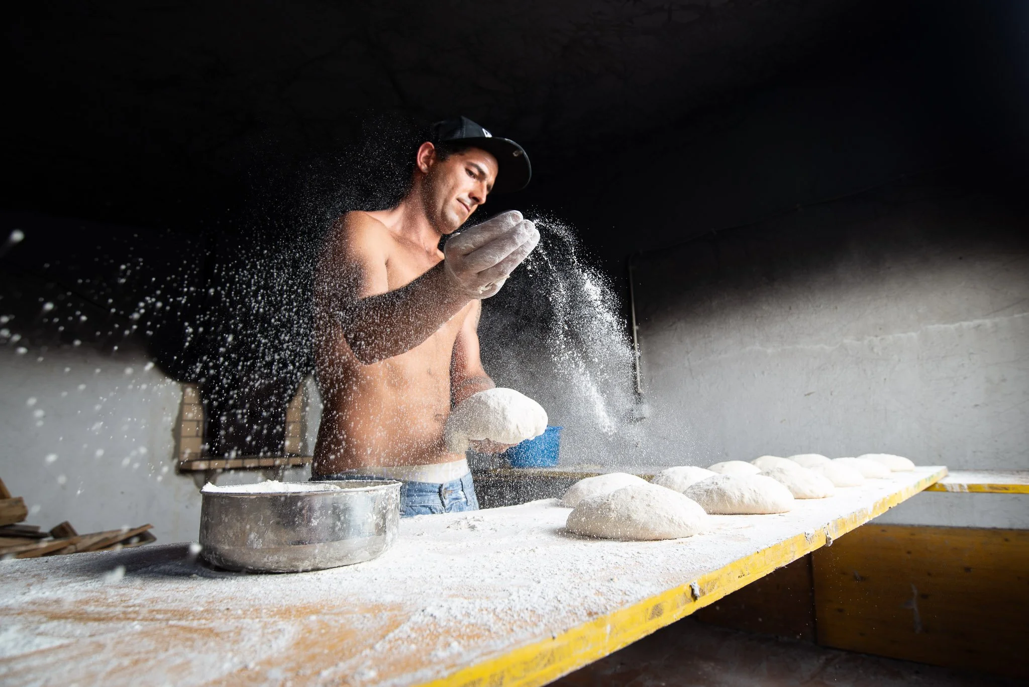 Man working with dough in a bakery, shirtless, wearing gloves, dusting flour on bread loaves on a wooden table.