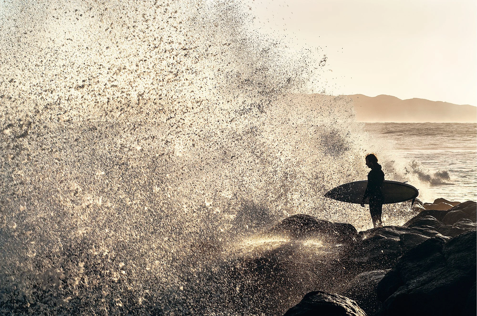 A surfer prepares to jump off the iconic 'Jump Rock' at Manu Bay during a heavy high-tide swell. Captured in the crisp morning light of Raglan, New Zealand.