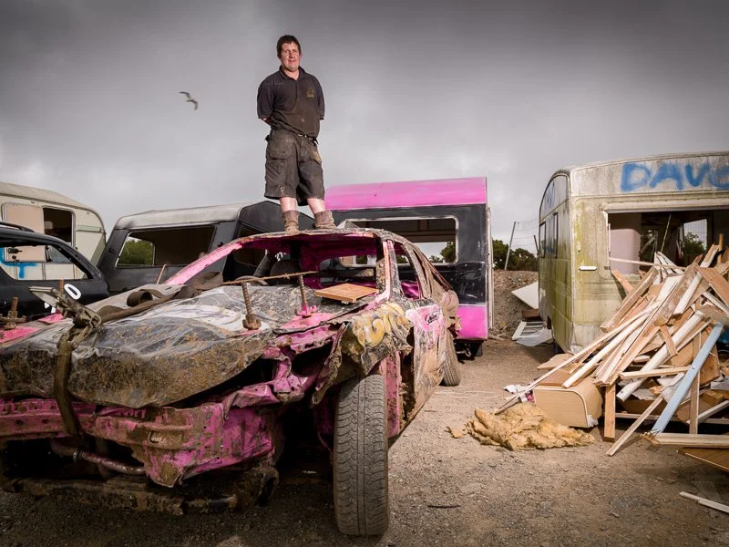 A man standing atop a heavily damaged pink and black car in a junkyard with other wrecked vehicles and debris around, under a cloudy sky.