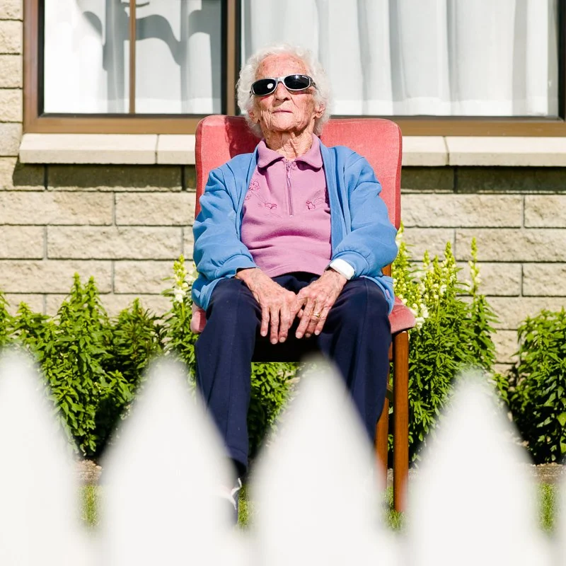 An elderly woman sitting outdoors on a pink chair, wearing sunglasses, a pink polo shirt, a blue jacket, dark pants, and white sneakers. She is behind a white picket fence with green plants in the background and a house with a beige brick wall and wi