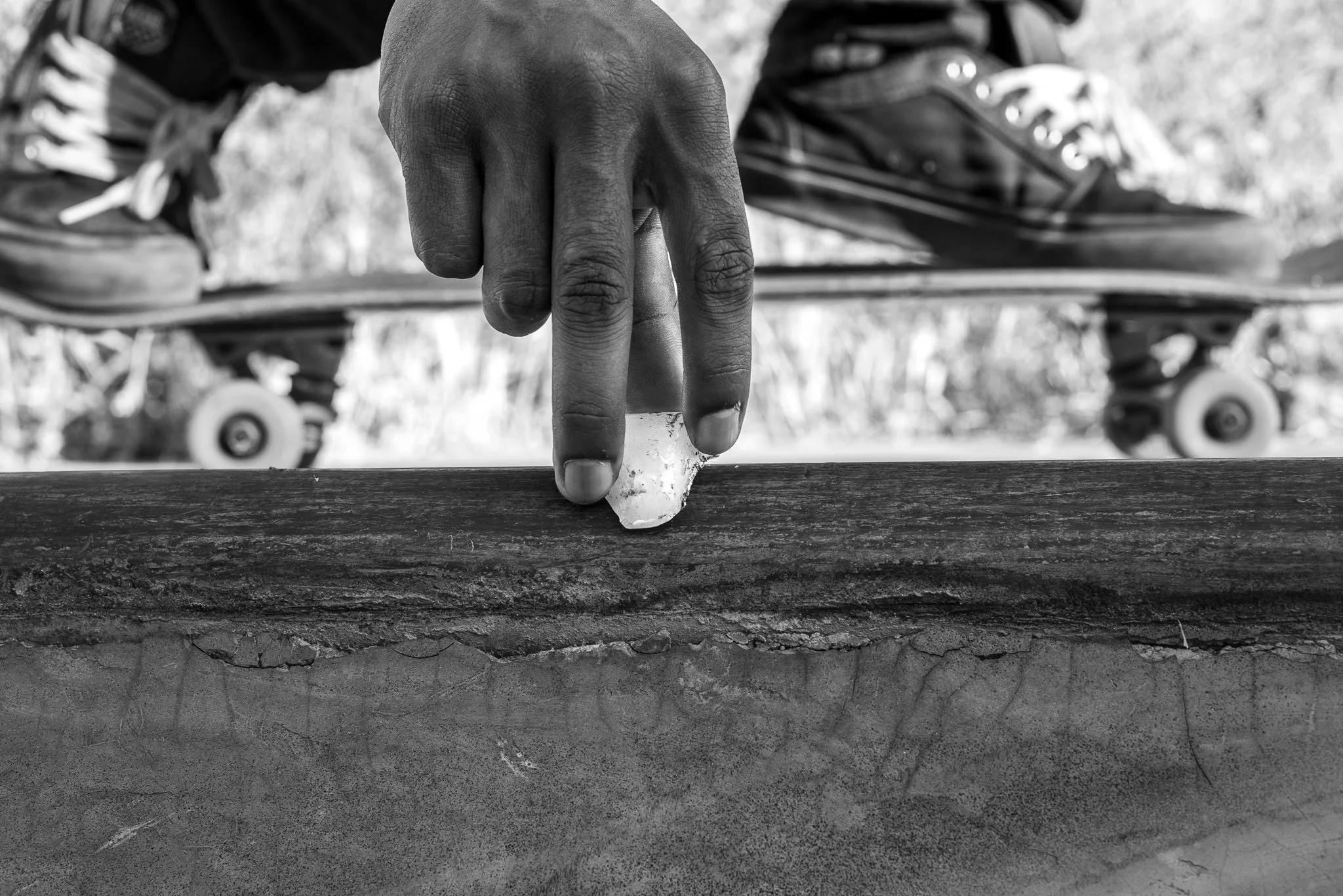 Close-up of a person's hand peeling a piece of ice off a wooden surface, with a skateboard and grass in the blurred background.