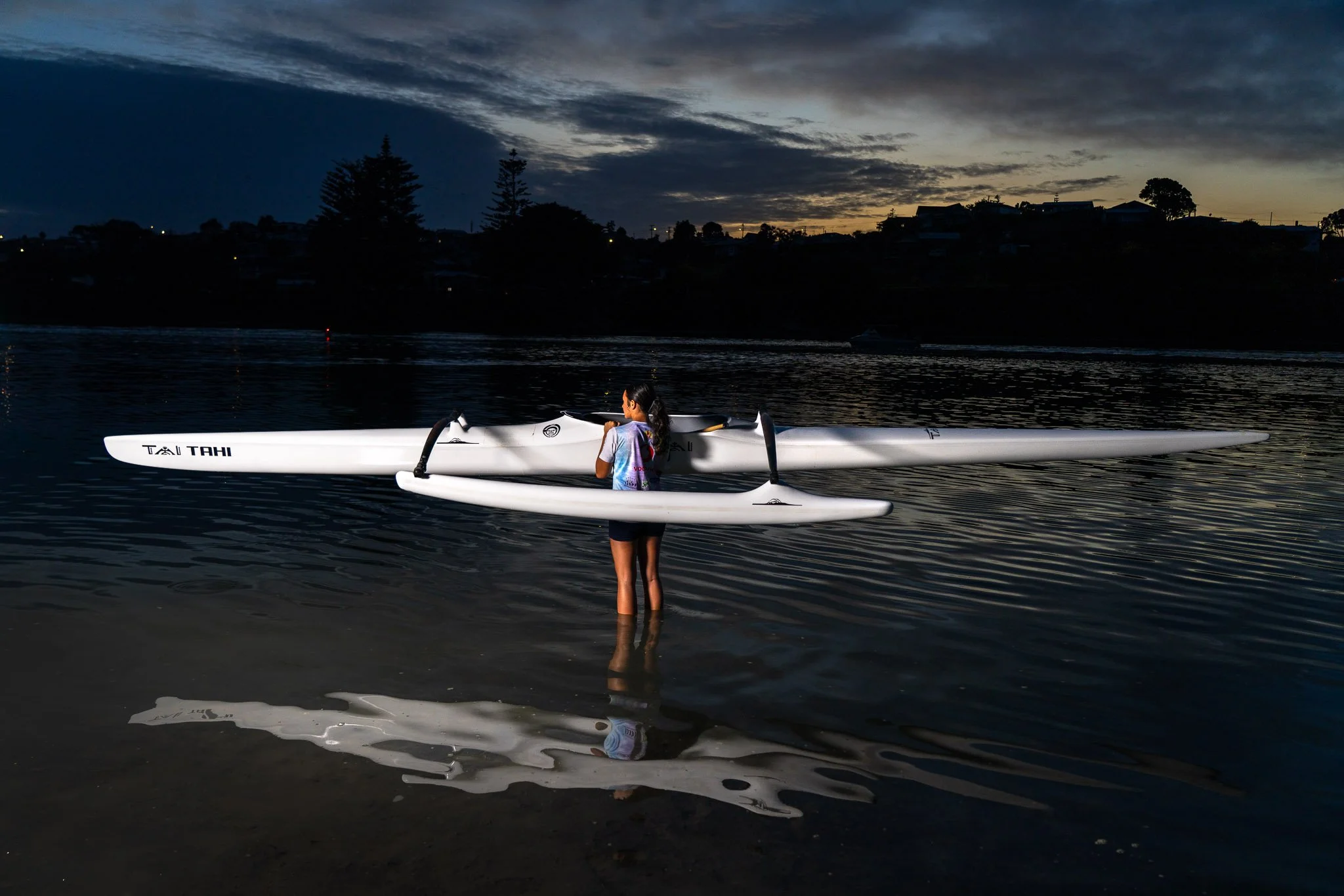 A girl standing in shallow water holding two kayaks at sunset or dusk.