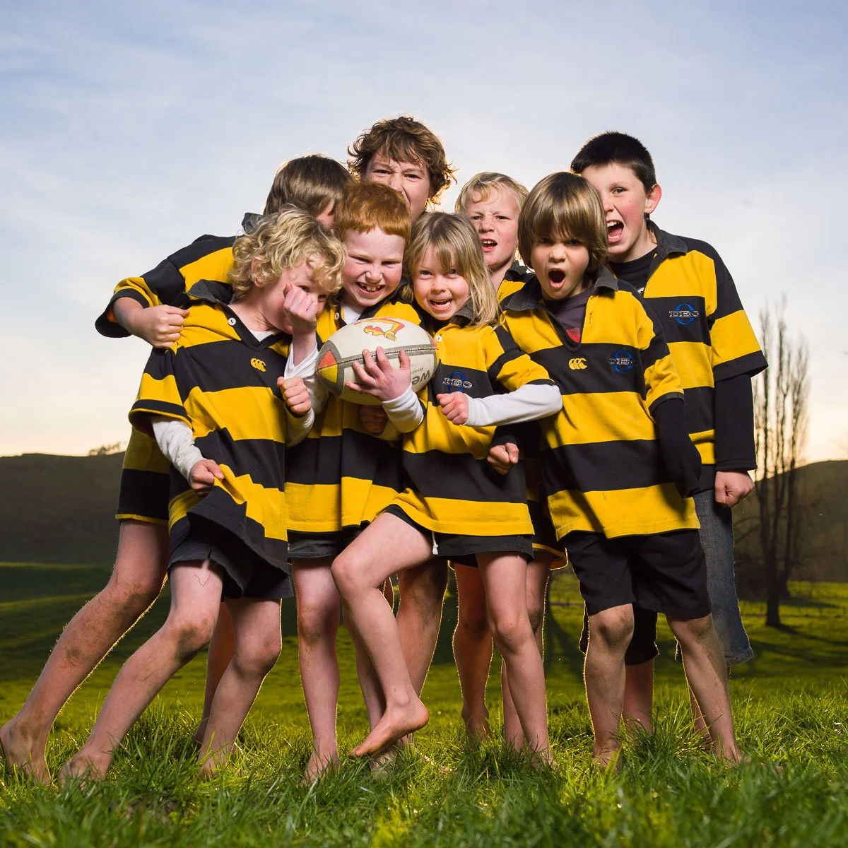 A group of children in rugby uniforms holding a rugby ball, outdoors on a grassy field during sunset.