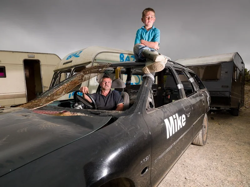 A young boy with arms crossed sitting on top of a black car labeled 'Mike', with a man sitting in the driver's seat. The car is parked outdoors with trailers in the background.
