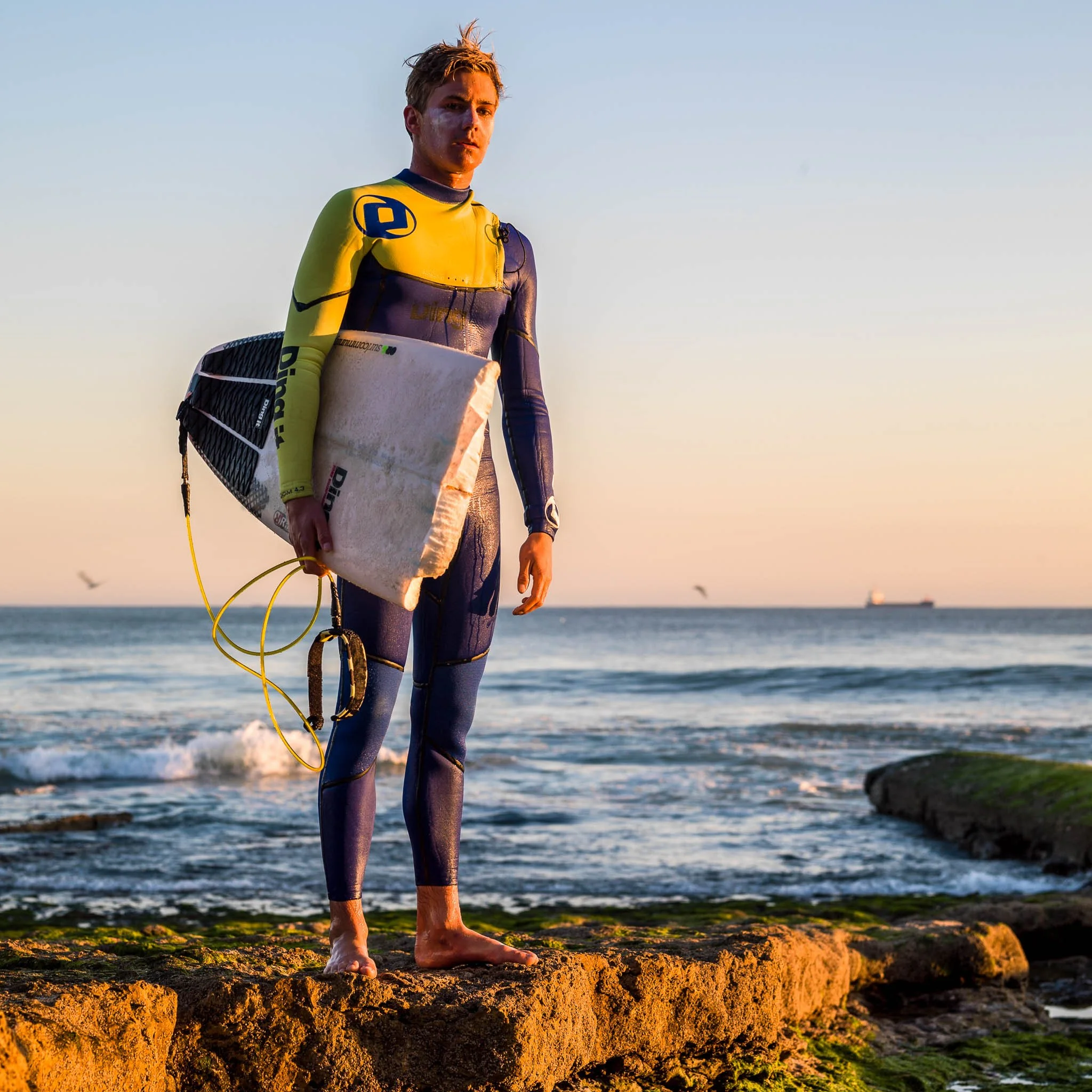 A young male surfer standing barefoot on a rocky beach at sunset, holding a surfboard, and dressed in a wetsuit.