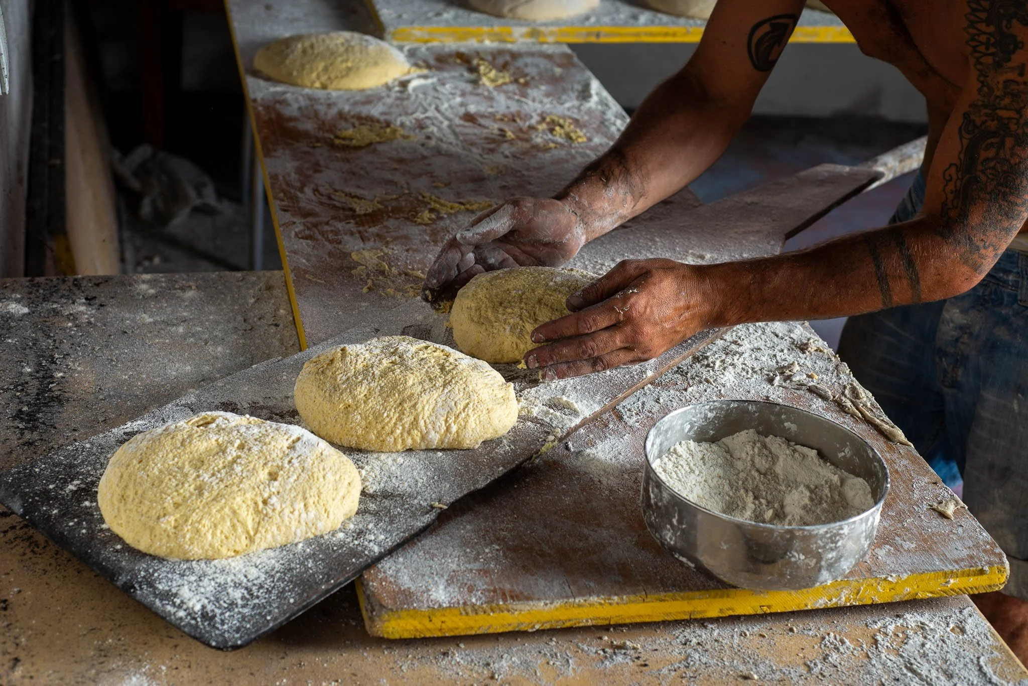 A person preparing dough on a floured surface with loaves of bread, a bowl of flour, and a mirror reflecting the hands working.