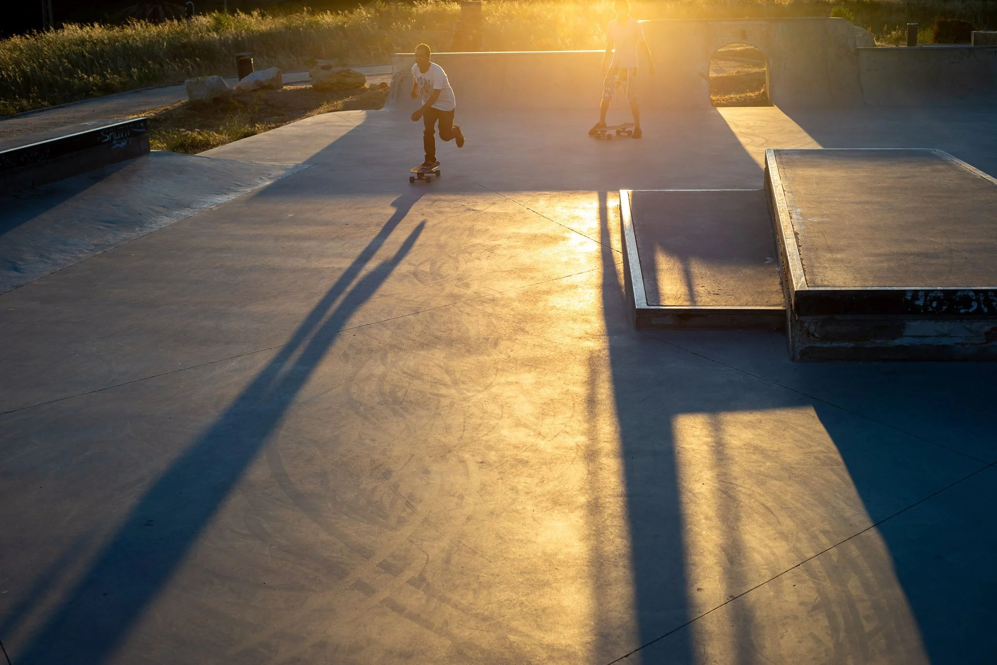 Two children skateboarding at a skate park during sunset, with long shadows cast on the concrete surface.