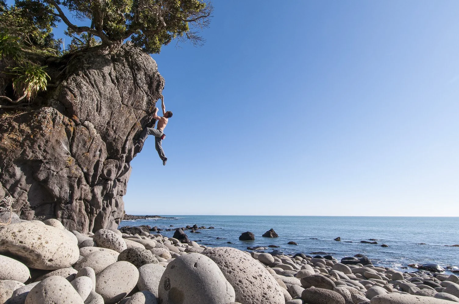 A person climbing a large rock formation on a rocky beach, with the ocean and a clear blue sky in the background.