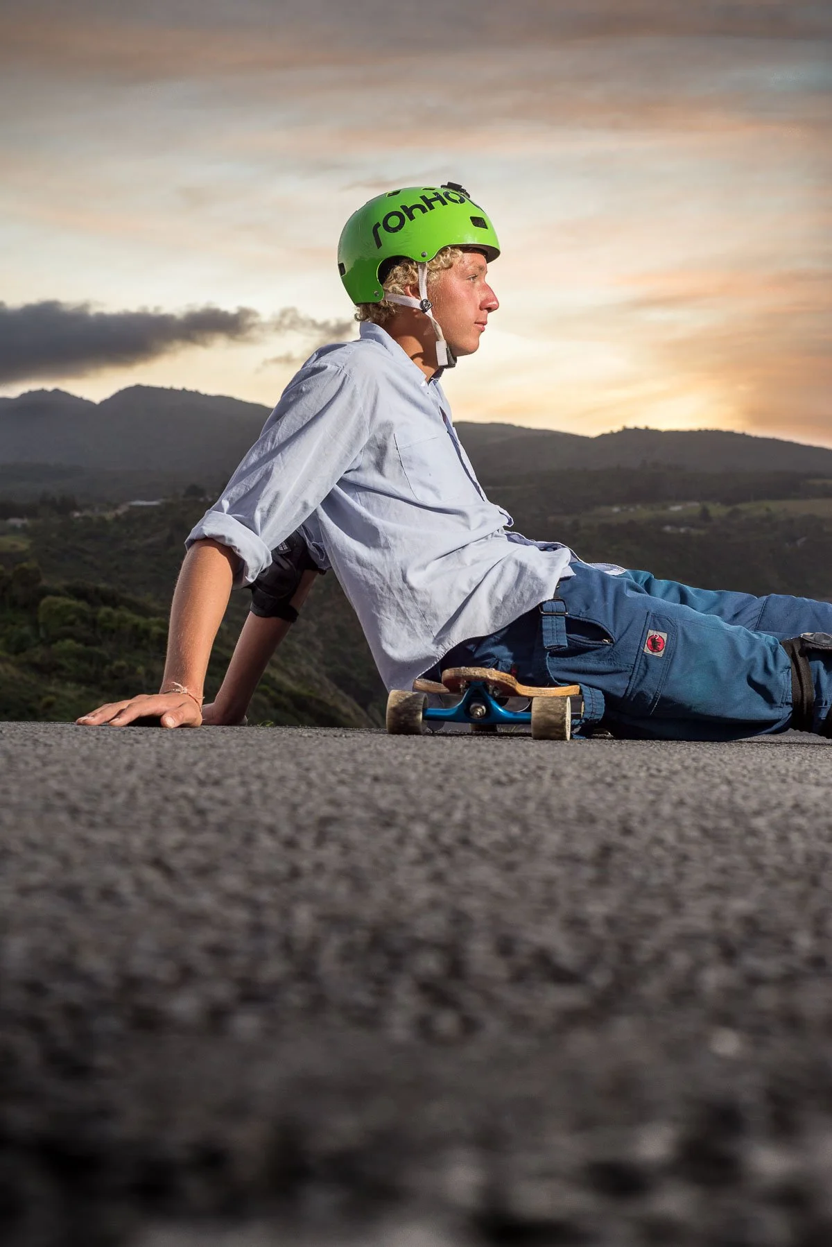 A young person wearing a green helmet and casual clothing, sitting on the ground with a skateboard beside them, in an outdoor landscape at sunset.