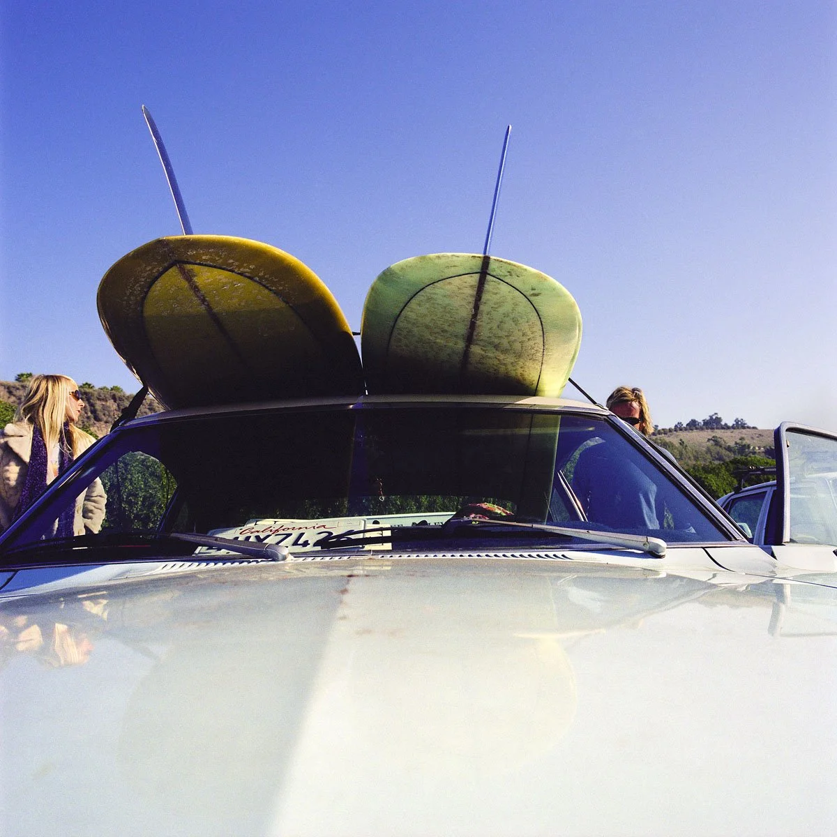 Professional surf lifestyle photography: A vintage car at Rincon, California, with two surfboards on the roof and a prominent 'California' number plate. A woman and a man stand beside the car. Shot by Paul Abbitt. 